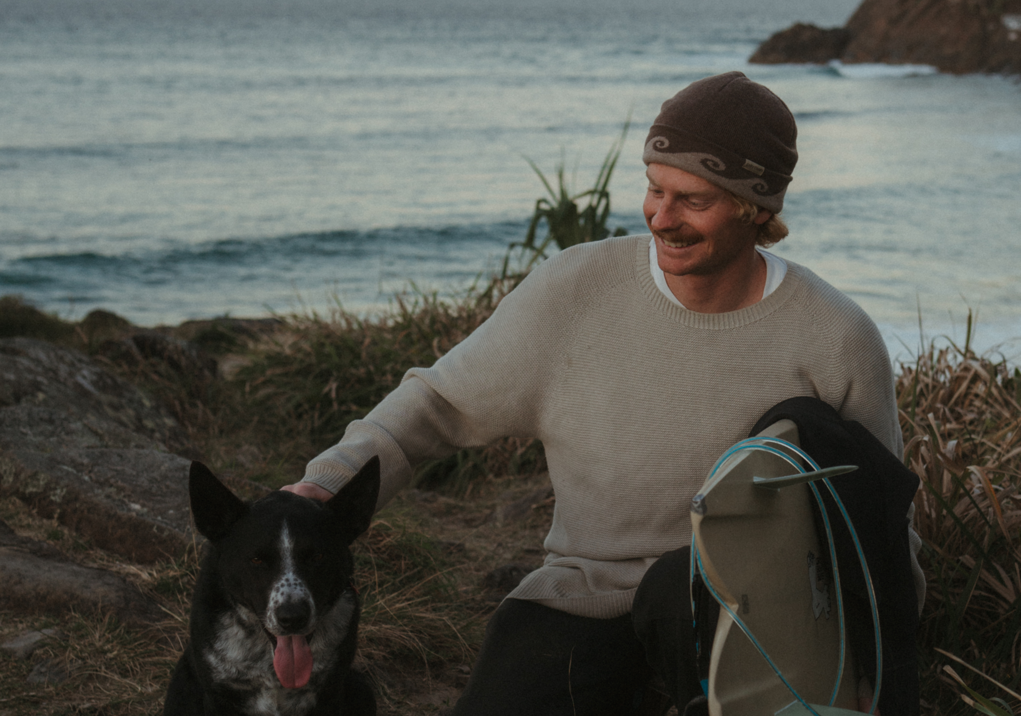 Man wearing a barrel beanie with a dog and surfboard sitting on a beach at sunset