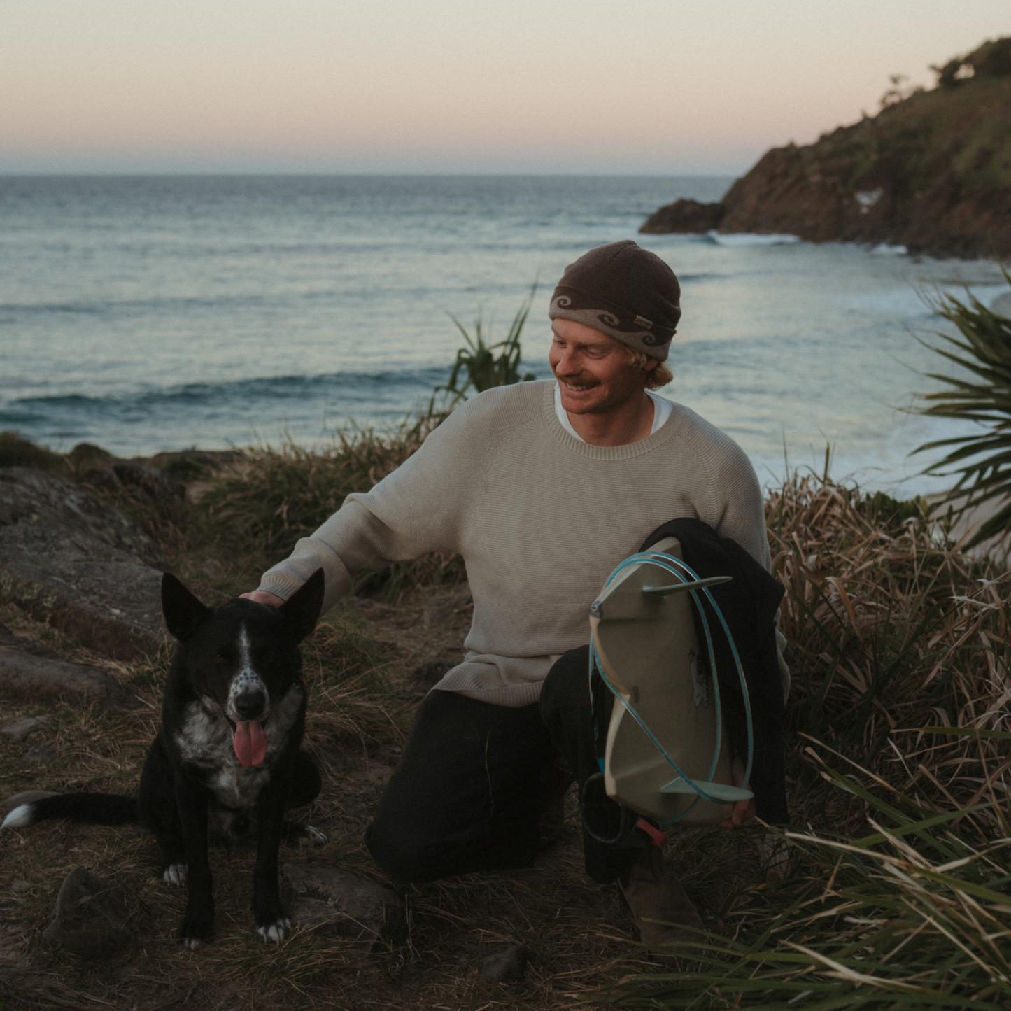 Man wearing a beanie with a dog and surfboard on a beach at sunset