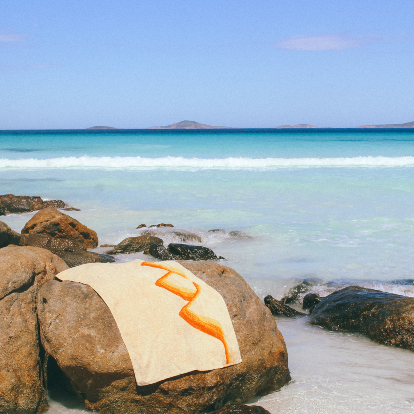The Classics Beach Towel is laying on a large rock with the ocean breaking around it.