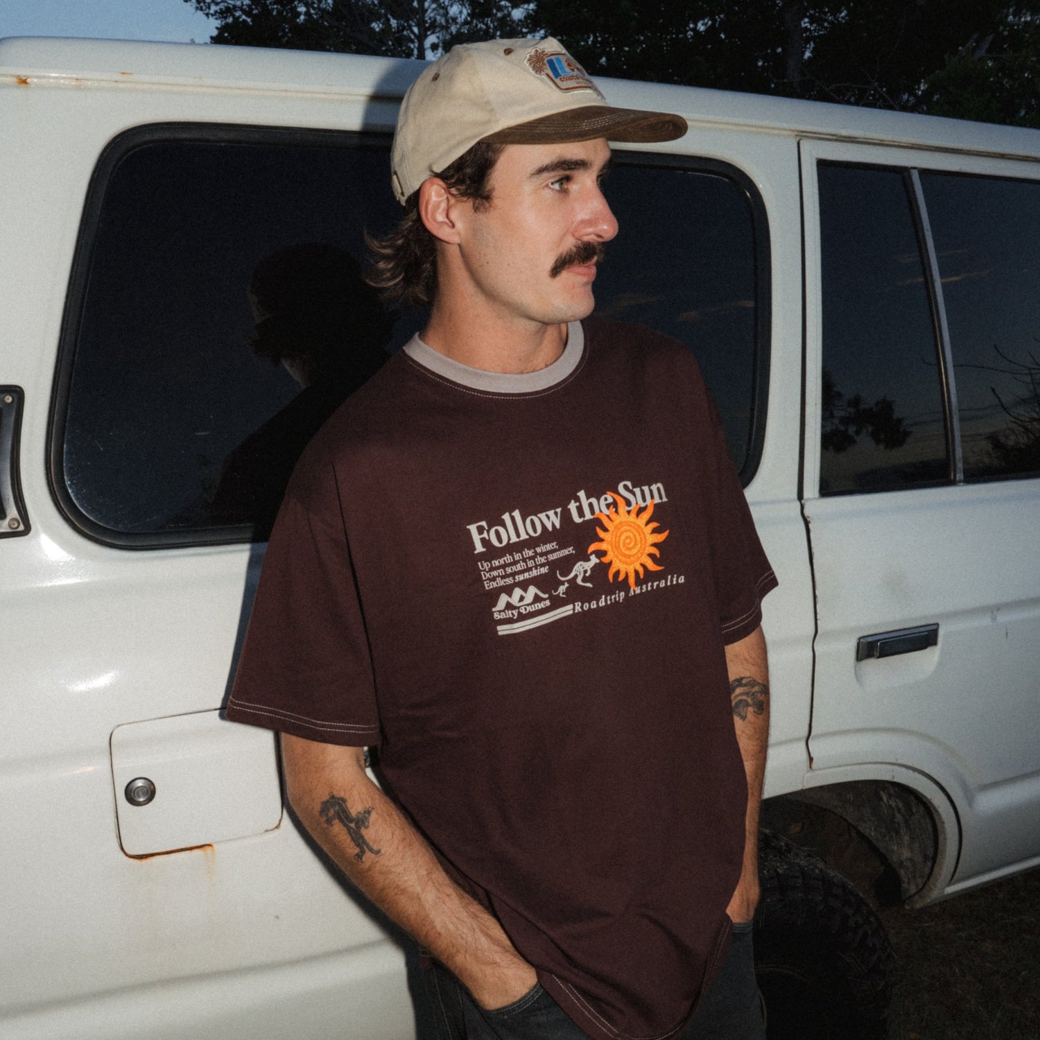 A man wearing a brown graphic t-shirt with 'Follow the Sun' printed on the front, paired with a beige 5-Panel cap, standing next to a white Landcruiser at dusk.