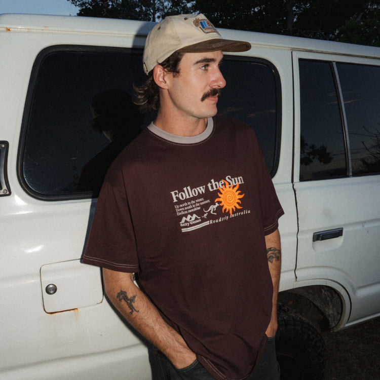 A man wearing a brown graphic t-shirt with 'Follow the Sun' printed on the front, paired with a beige 5-Panel cap, standing next to a white Landcruiser at dusk.