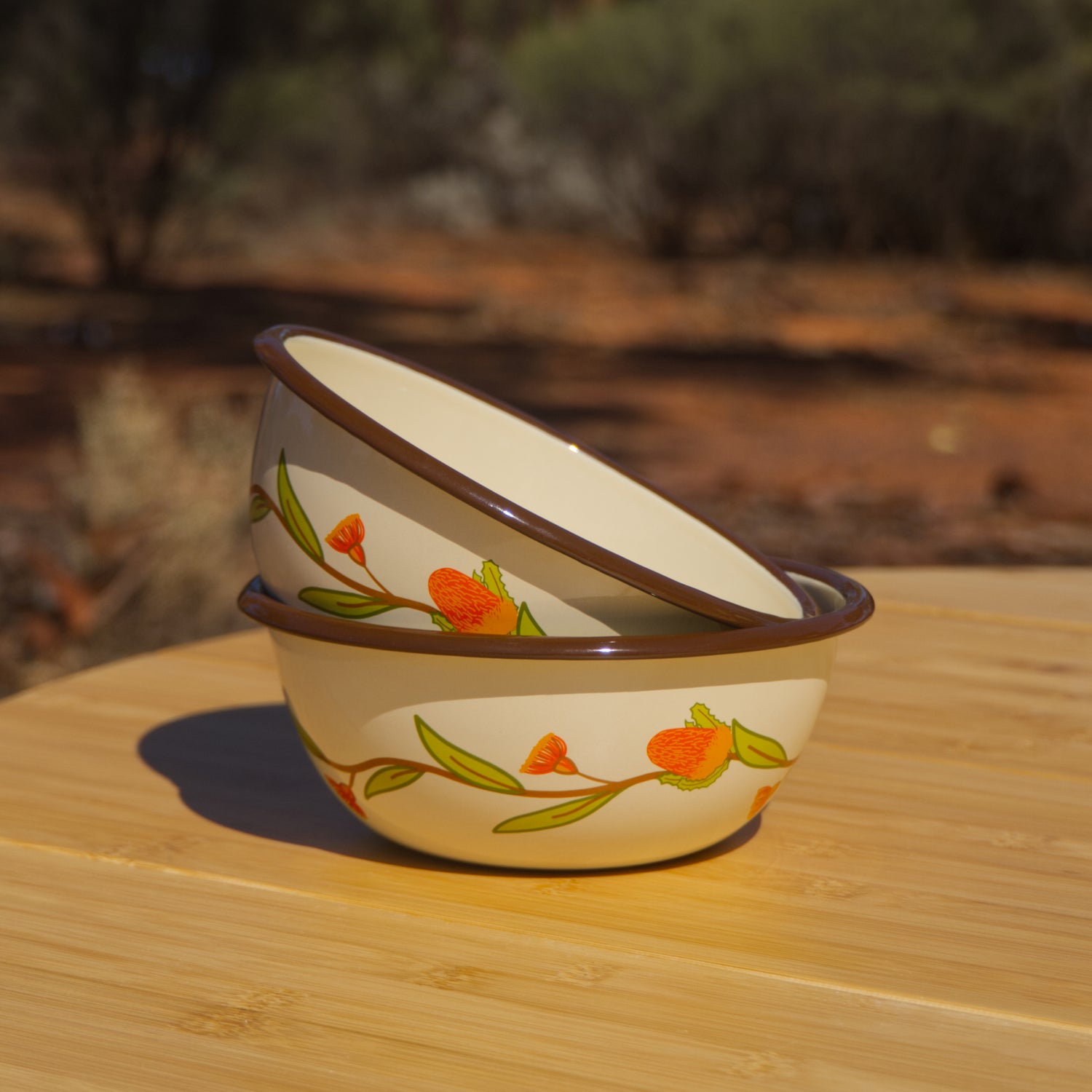 Two cream and brown coloured enamel bowls stacked on a wooden table in the outback. The bowls feature an Australian Natives inspired floral design around the outside