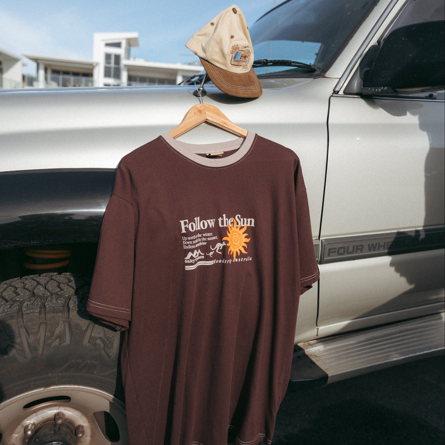 A brown graphic t-shirt hanging on a wooden hanger from a vehicle door, with a tan baseball cap resting on the hood. The shirt features a white graphic that reads 'Follow the Sun' with a sun and kangaroo illustration.