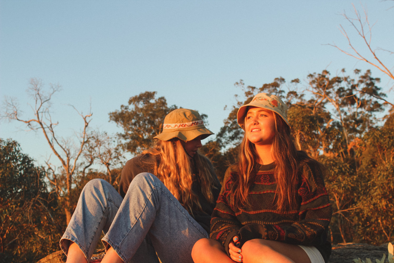 Two girls wearing the Australian Natives Hemp bucket hat and the classics hemp wide brim both looking out on a sunset in the bush