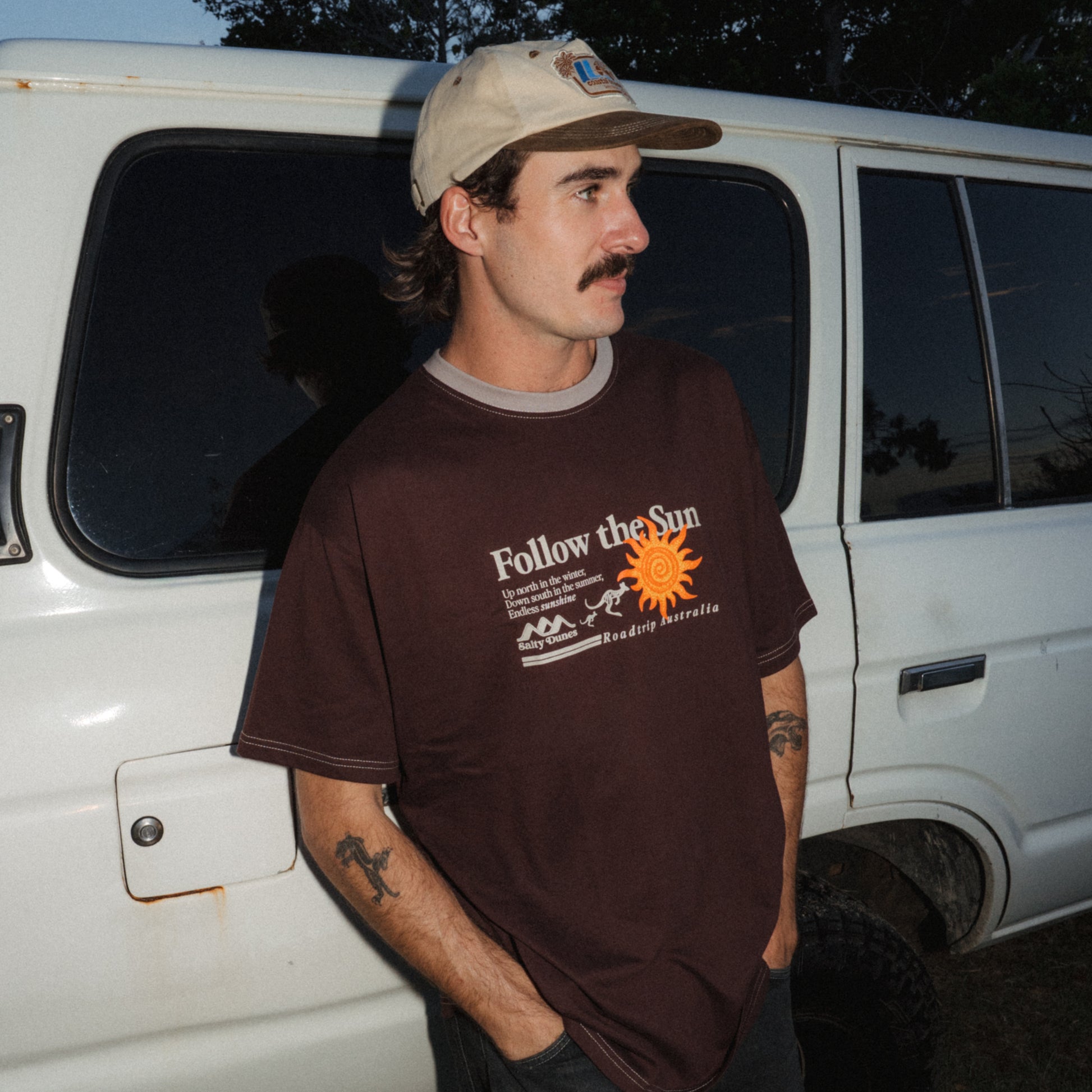 A man wearing a brown graphic t-shirt with 'Follow the Sun' printed on the front, paired with a beige 5-Panel cap, standing next to a white Landcruiser at dusk.