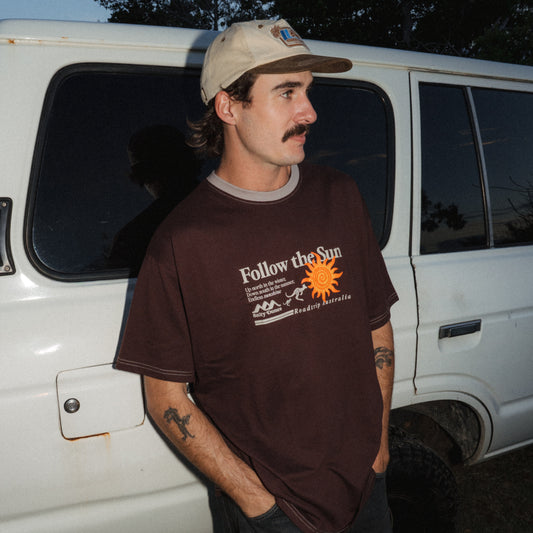 A man wearing a brown graphic t-shirt with 'Follow the Sun' printed on the front, paired with a beige 5-Panel cap, standing next to a white Landcruiser at dusk.
