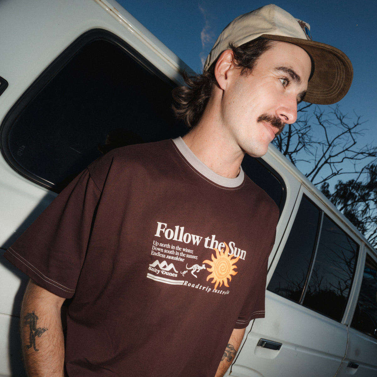 A man wearing a brown graphic t-shirt with 'Follow the Sun' printed on the front, paired with a beige cap, standing next to a white vehicle at dusk.