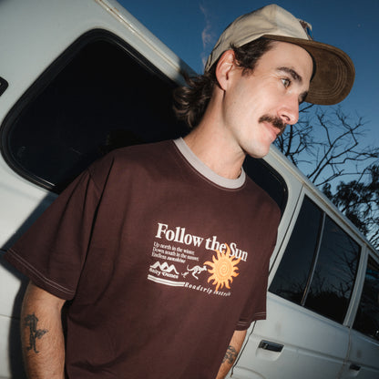 A man wearing a brown graphic t-shirt with 'Follow the Sun' printed on the front, paired with a beige cap, standing next to a white vehicle at dusk.