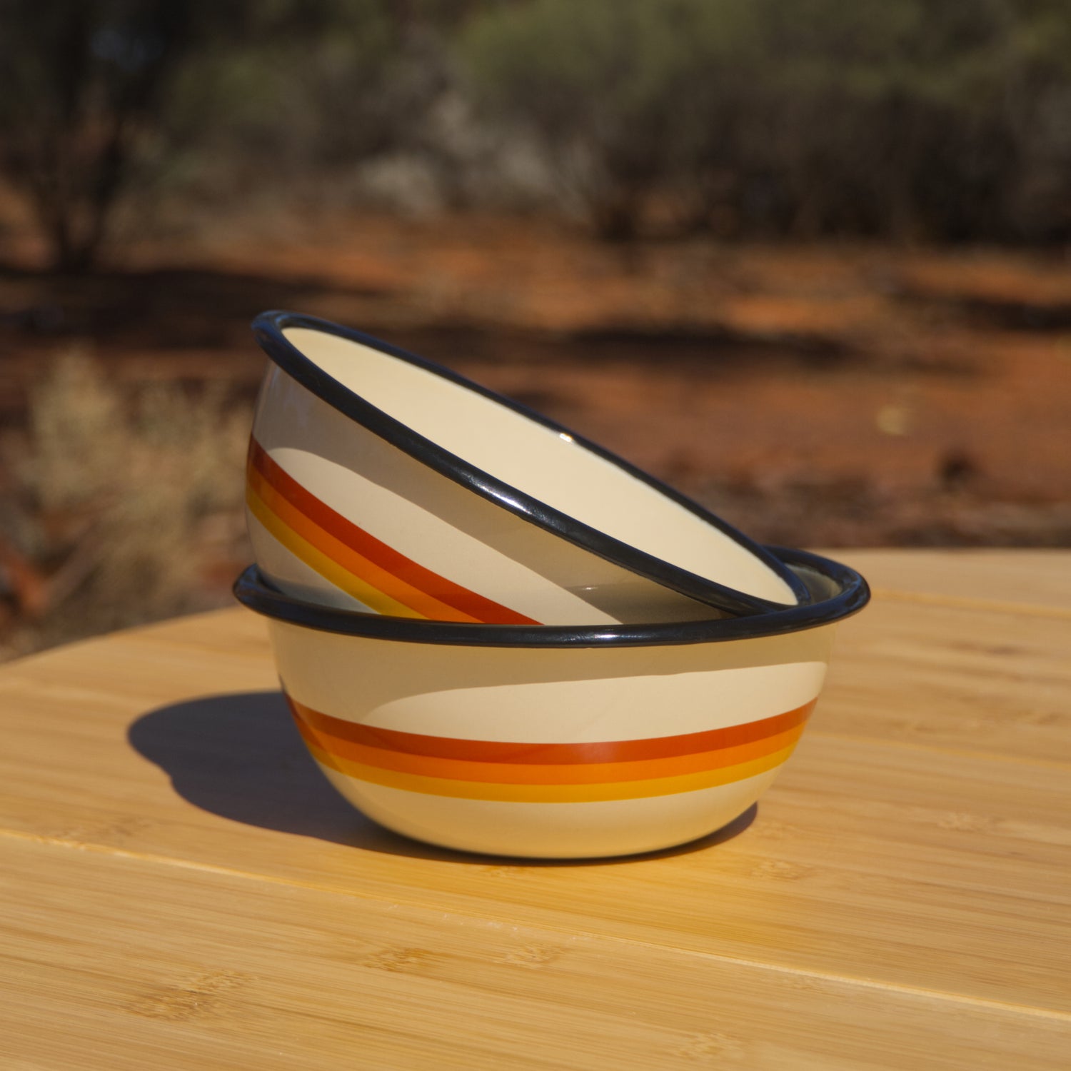 Stack of enamel bowls with a striped red, orange and yellow design wrapped around the outside. The stack is sitting on a wooden table in the outback.