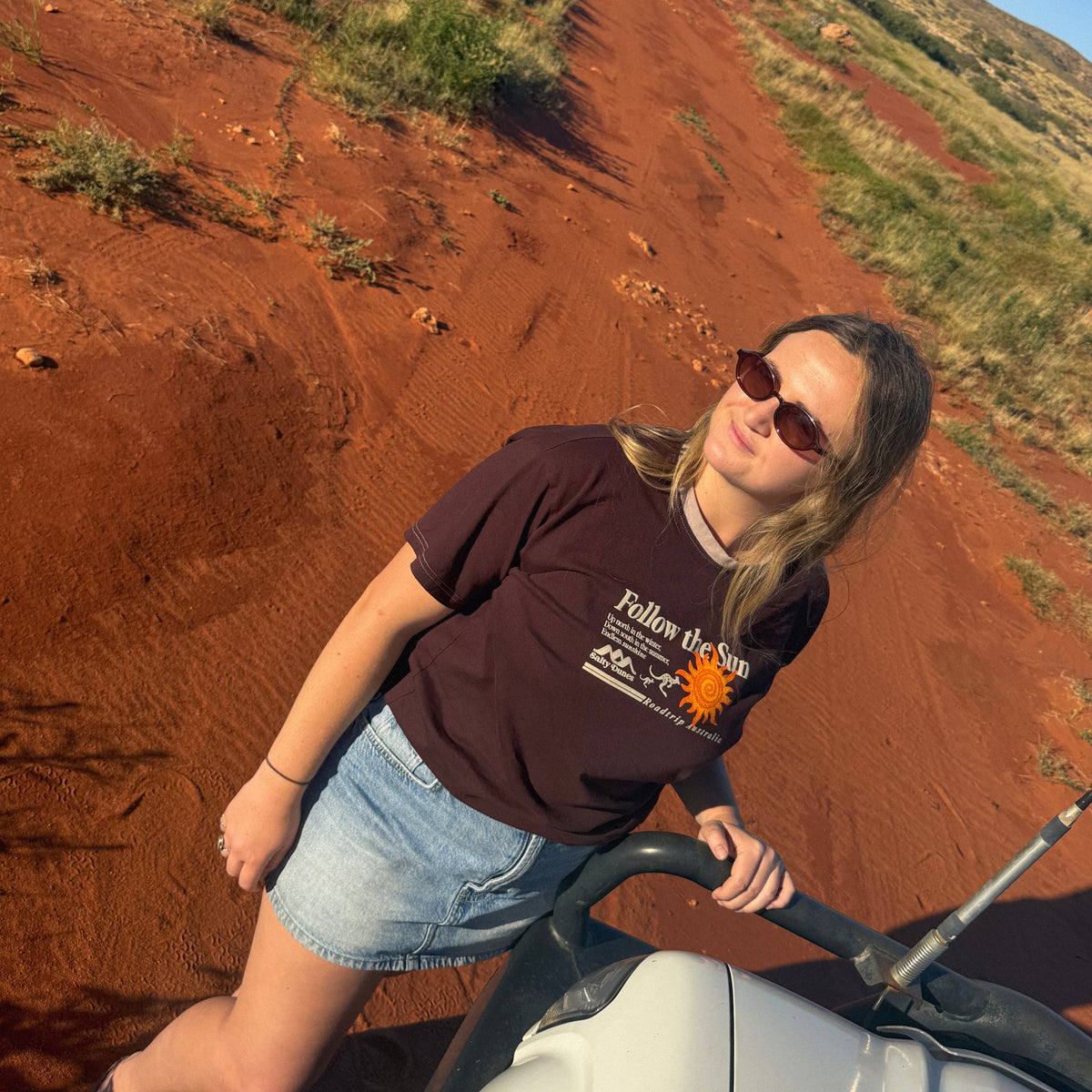 A woman wearing a dark brown graphic t-shirt and denim shorts holds a white hilux on a red dirt road in an outdoor australian landscape.