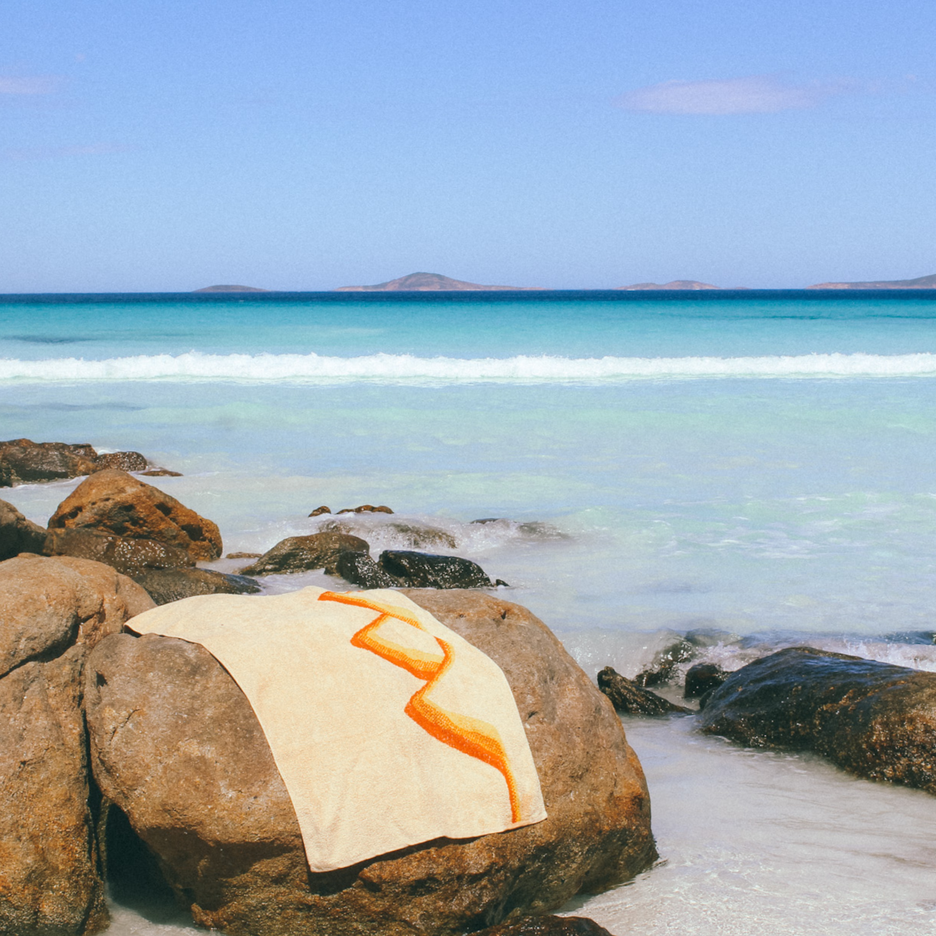 The Classics Beach Towel is laying on a large rock with the ocean breaking around it. 