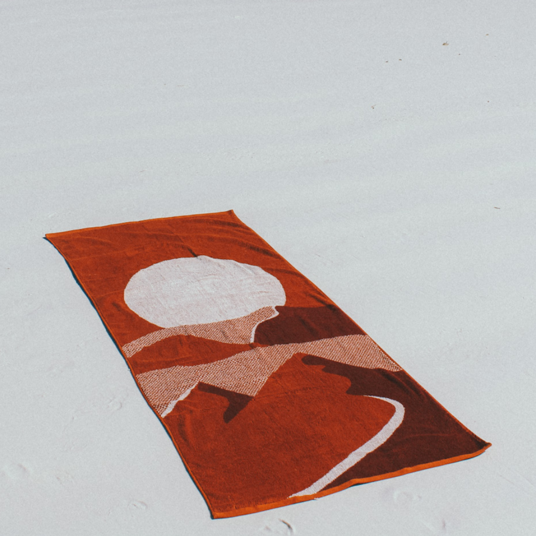 The Dunes Beach Towel lying down on a white sandy beach. You can only just see the start of the ocean at the top of the image