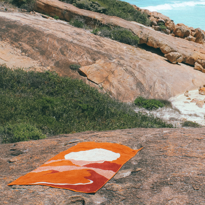 The Dunes Beach Towel, lying down on a large granite rock. The Beach is in the background where you can see rolling waves.