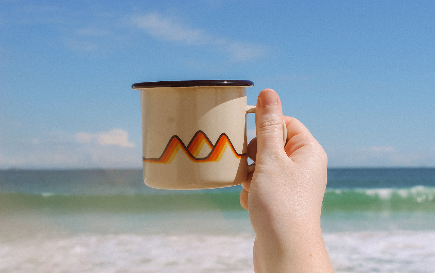 Someone holding a Salty Dunes classics enamel camp mug with the ocean in the background